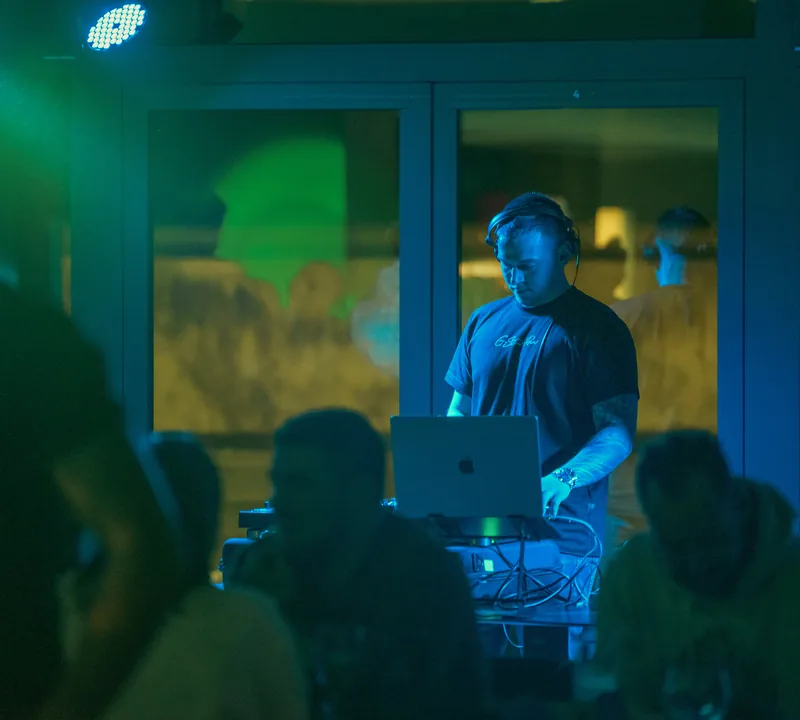 DJ a pultnál fejhallgatóval, laptoppal — zöld fény világítja meg a setet.